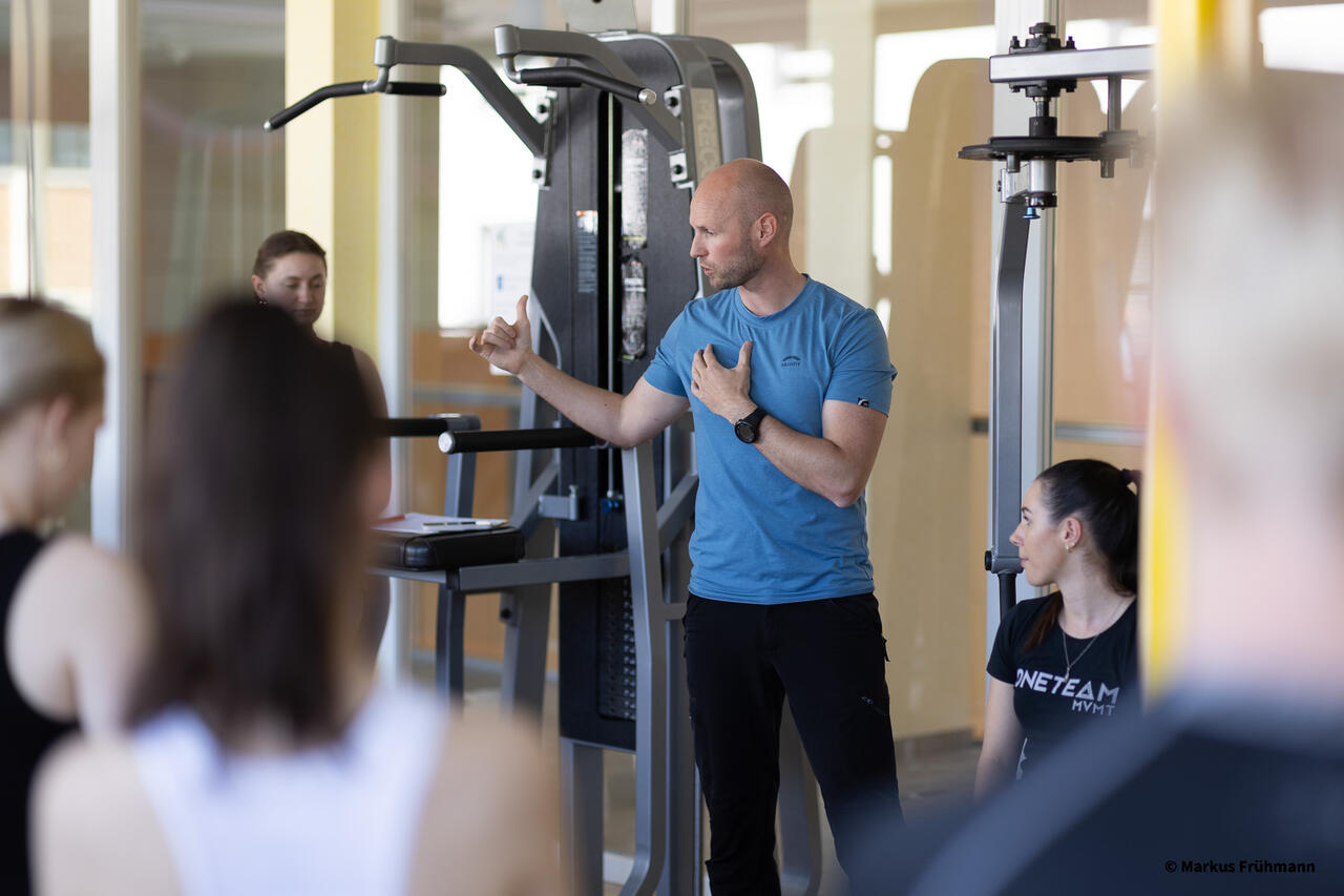 Traniner erklärt im Fitnessstudio vor einem Trainingsgerät.