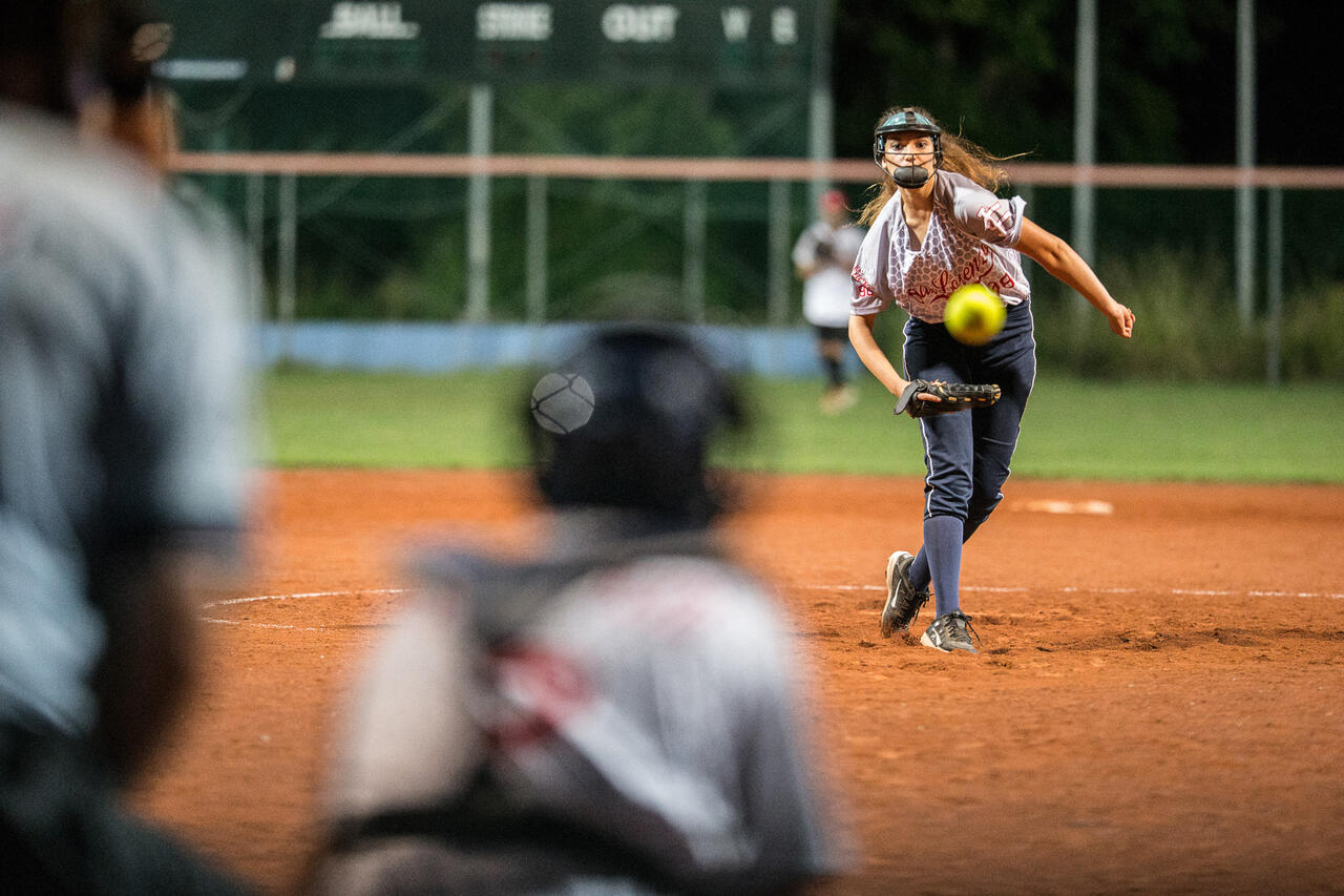 Eine Person mit Helm und Wurfhandschuh, der Ball fliegt in der Luft, zwei Personen erwarten ihn.