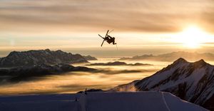 Bergkulisse bei Sonnenuntergang, im Vordergrund ein Ski Freestyler bei einem Sprung über einen Kicker