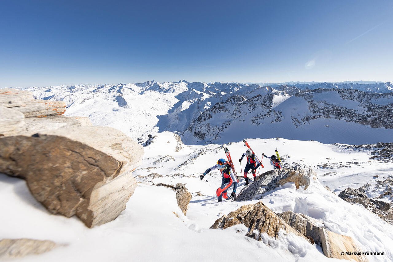 Drei Personen mit Ski am Rücken beim Aufstieg auf einen Schneeberg.