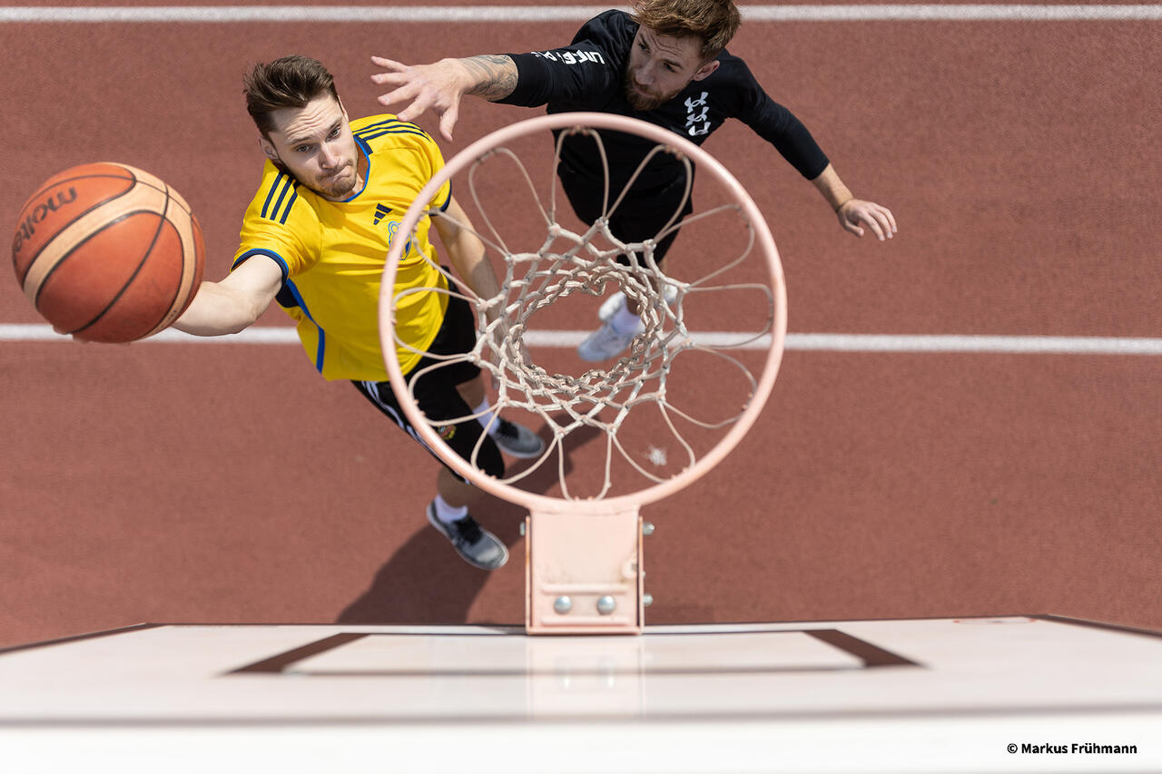 Von oben sieht man eine Person mit Ball in der Hand in Richtung Korb unterwegs, die zweite Person versucht noch zu verteidigen.