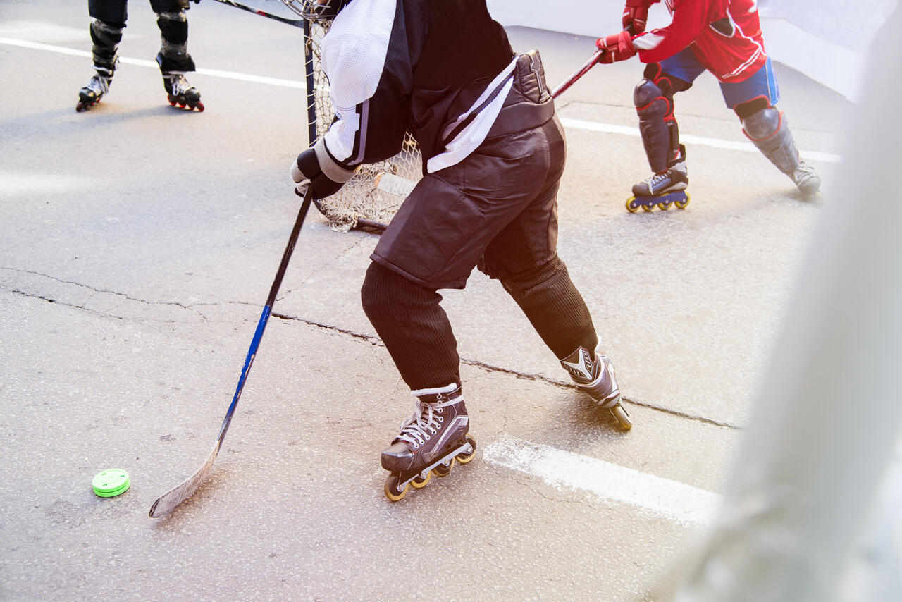 Ausschnitt einer Person mit Schläger, Ball und Skates, weitere Personen daneben.