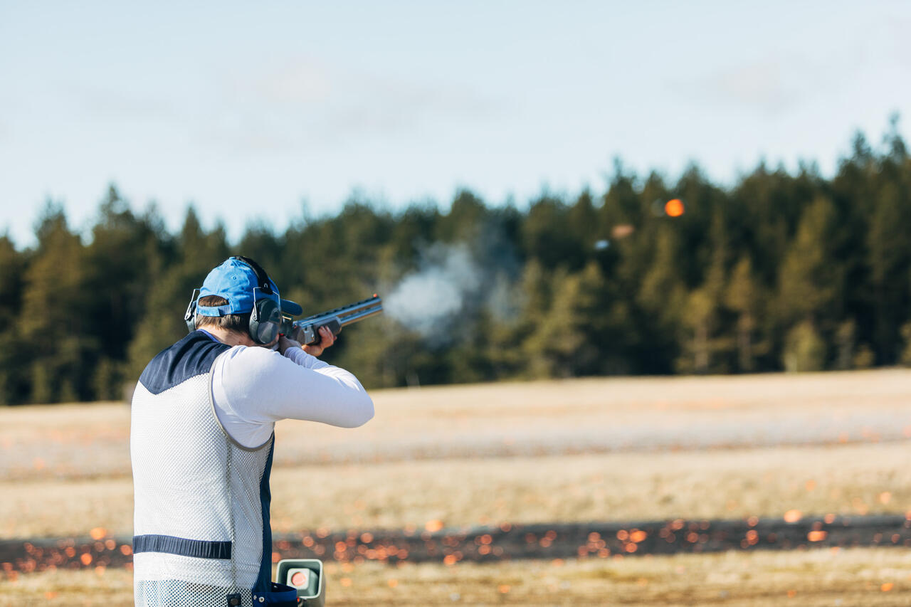 Ausschnitt einer Person von hinten mit Gewehr in der Hand und Ohrenschutz kurz nach Schuss, Rauch liegt in der Luft.